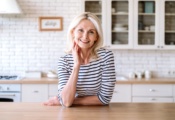 Cheerful middle aged woman standing near table at home kitchen, smile and looking at camera. Portrait of confident joyful female entrepreneur in casual clothes
